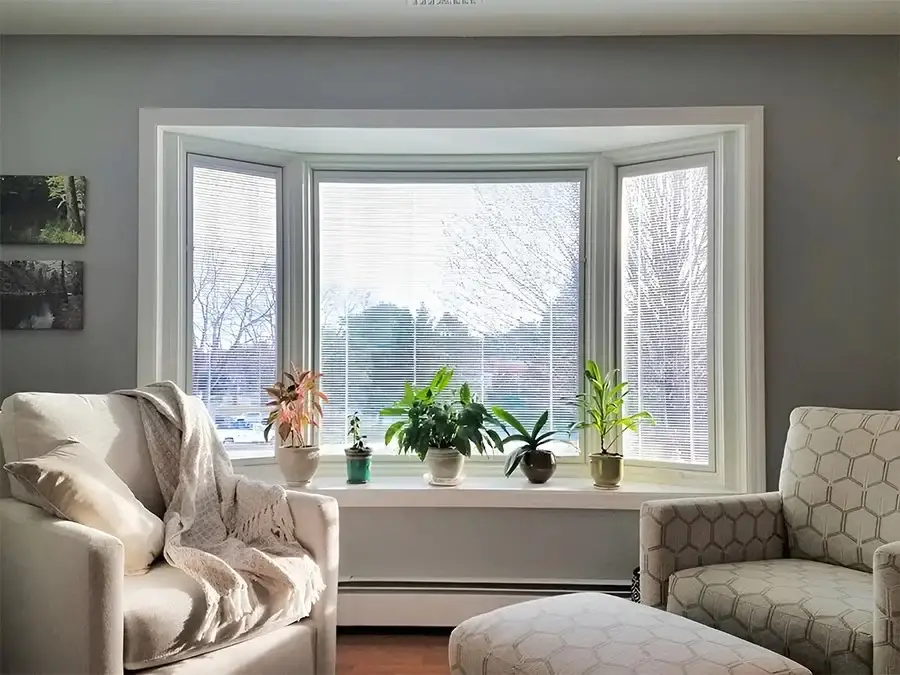A set of three windows, bay style, in a cozy living area with built-in blinds and white exterior wood clad framing. In front of the window is a set of plants in small planters on the shelf. There are two sitting chairs visible with lots of sunlight coming into the room.