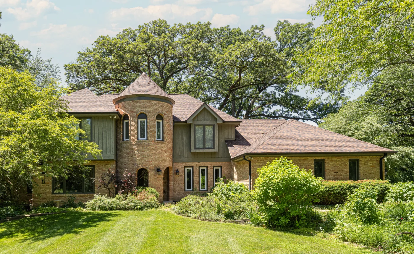 A beautiful, large, wide, luxurious, soft brown brick home in the Chicagoland area with a manicured lawn and lush landscaping. It’s almost half craftsman and half Victorian with a prominent two-story rounded tower, complex architecture, new shingle roofing, and various picture windows with white and brown framing.