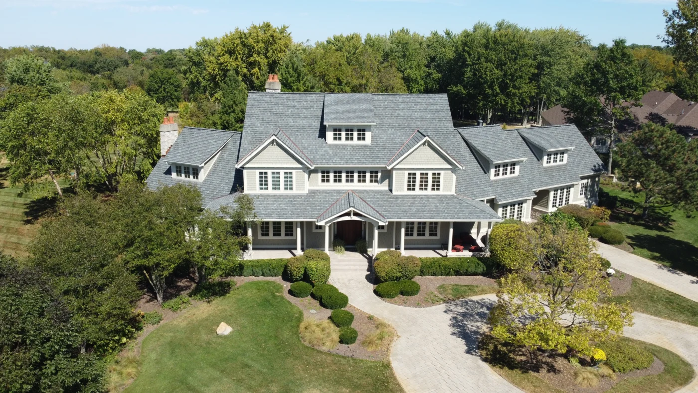 Better, More Effective Residential & Commercial Exterior Remodeling Systems An aerial shot of a large Chicagoland home with many sets of three white windows with traditional grids of different sizes, a new asphalt shingle roofing system installed by Apex Exteriors, and new light colored fiber cement siding and luxurious, curated landscaping.
