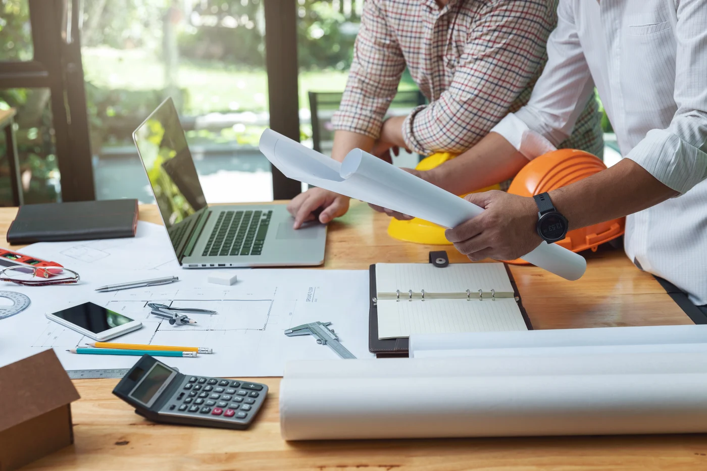 Construction professionals reviewing building plans and architectural drawings at a work table. Rolled blueprints, drafting tools, hard hats, a laptop, and calculator are visible, illustrating careful planning, material coordination, and detailed preparation for a commercial new construction project.