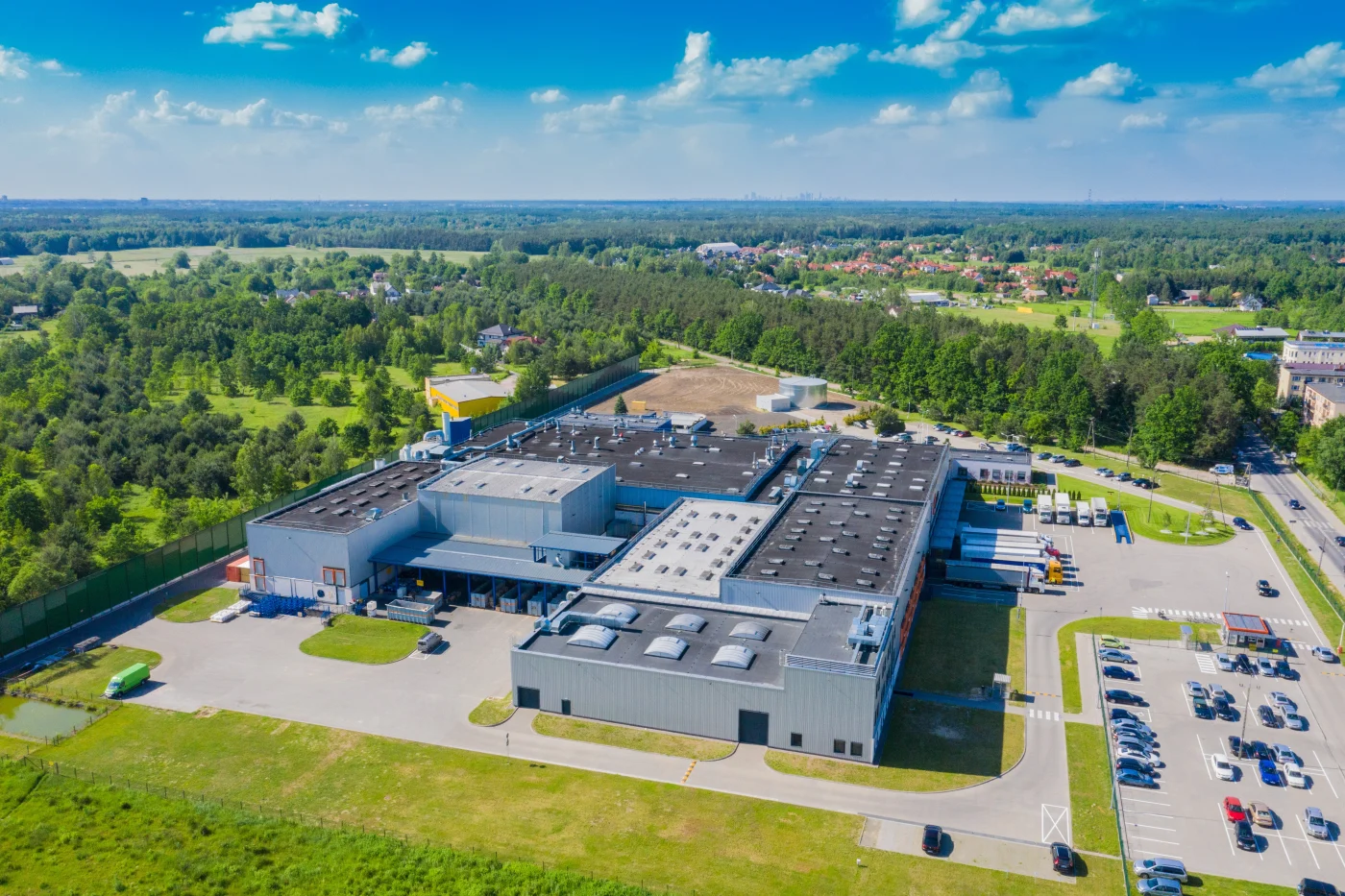 Aerial Drone View On Distribution Center. Logistic And Transport Concept An aerial view of a large industrial facility with light gray metal siding and multiple flat rooftops, surrounded by parking lots, semi-truck docks, and green open space. The property is located in a semi-rural area with trees, fields, and distant residential neighborhoods.
