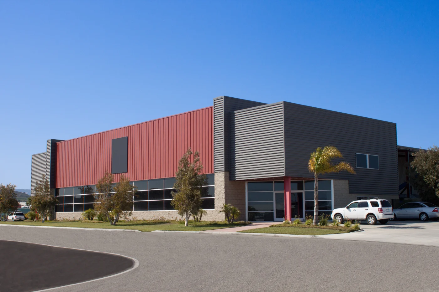 Commercial Siding Repair Protects Prevents Larger, Costly Issues A modern, two-story industrial or commercial building featuring dark gray horizontal metal siding on one side and vertical red metal panels on the other. The ground level includes large windows and a stone veneer base. A few parked vehicles and palm trees are visible in the foreground.