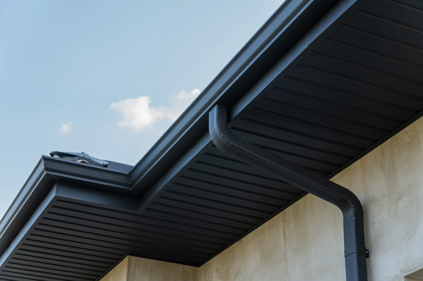 A close-up view of a modern black gutter and downspout system installed on a multi-family building. The structure features a smooth, light-colored stucco exterior and dark soffit panels under the overhang. The clean, angular lines of the roof edge and gutter system emphasize professional installation and low-maintenance materials.