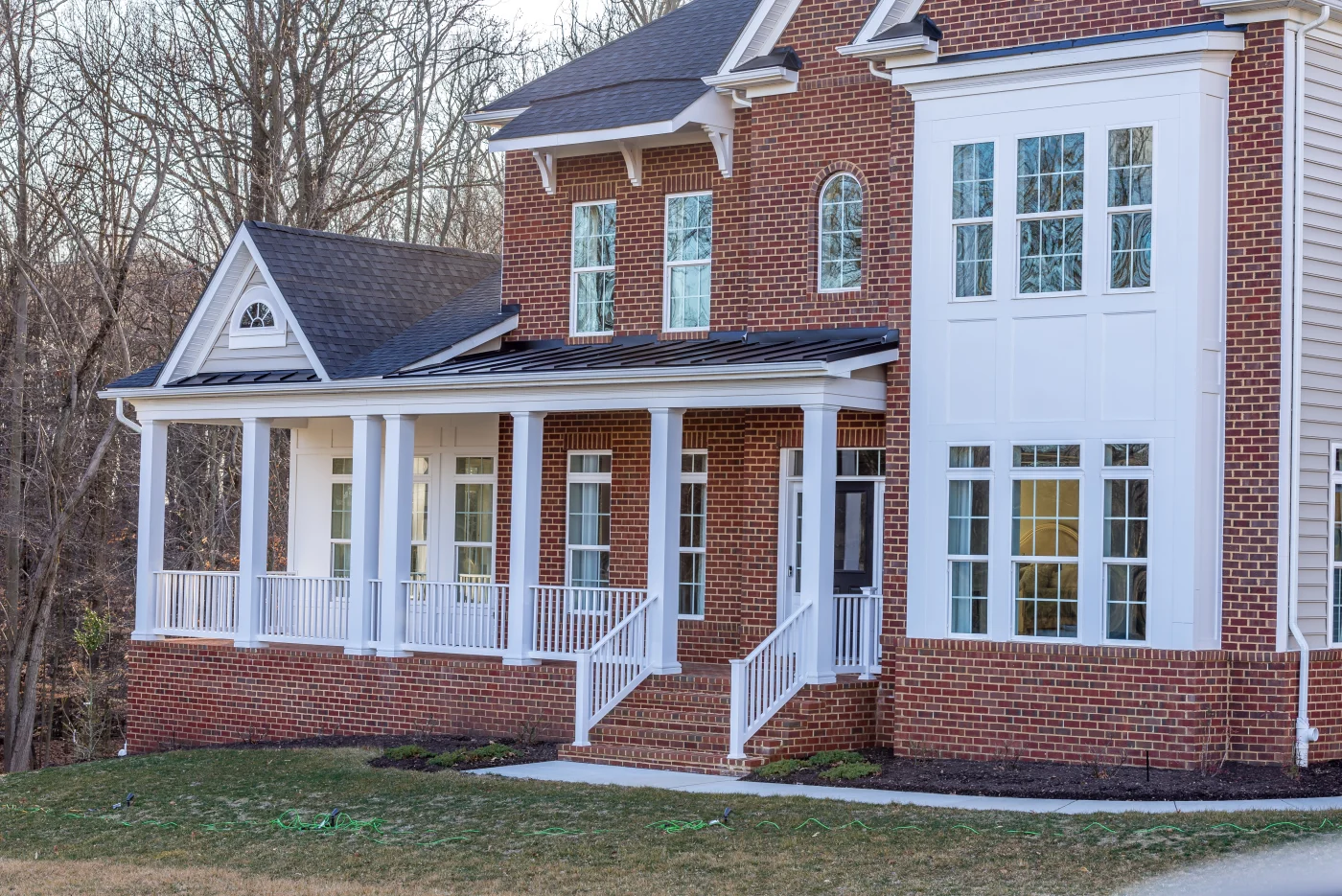 Apex Exteriors New Home Fascia Services In The Chicagoland Area A large, luxurious two-story red brick home with a section of white siding going up both sections. The windows are all white with traditional grids – mainly double-hung. It has a covered porch area with white pillars and railing and metal roofing with snow guards. White soffit and fascia. Shingle roofing.