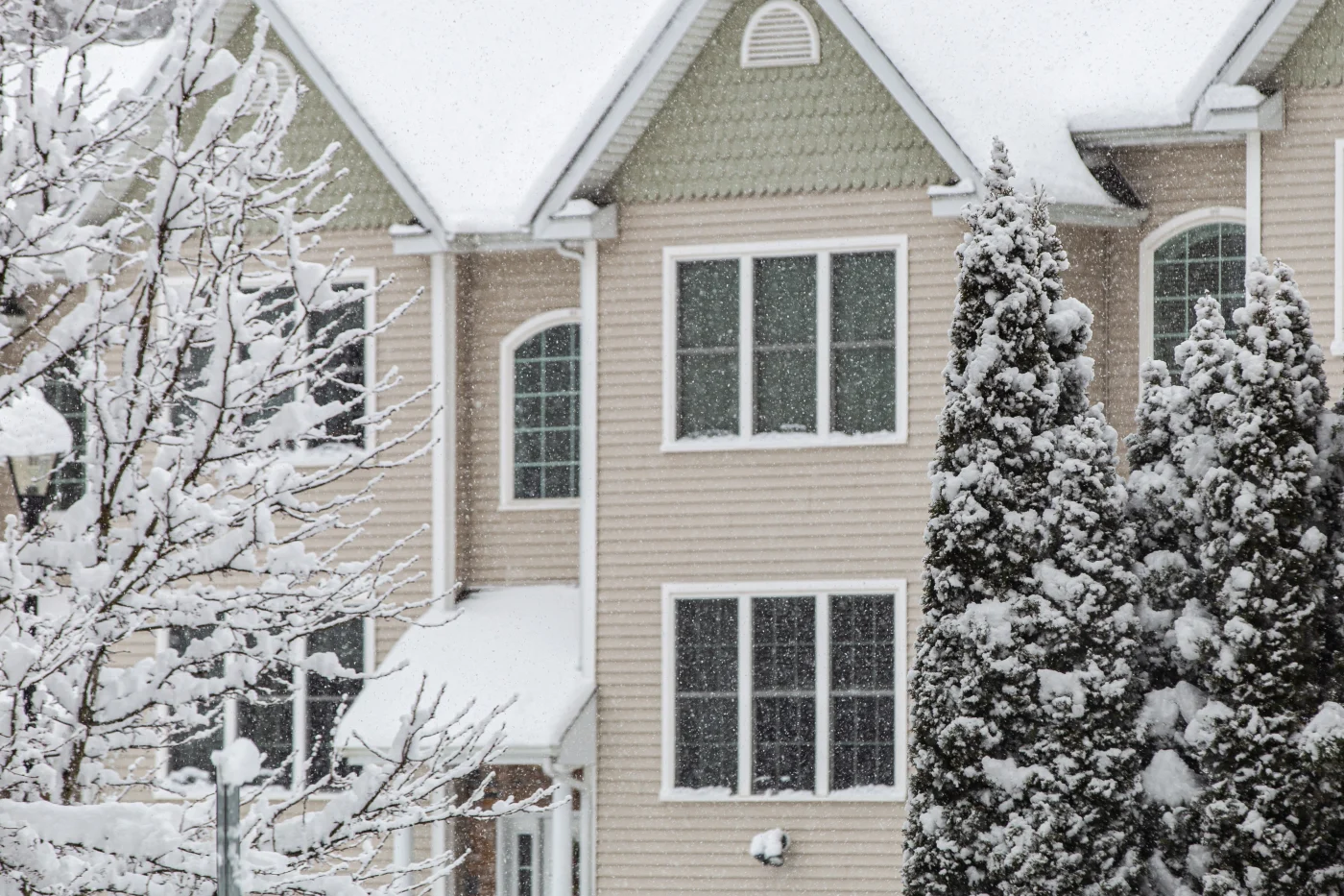 Apex Exteriors New Home Siding Services In The Chicagoland Area It’s snowing. There are trees almost covered in it. Between the trees is a large residential structure with beige horizontal panel siding around the windows, and more decorative siding around roof venting, white fascia, soffit, and window frames.