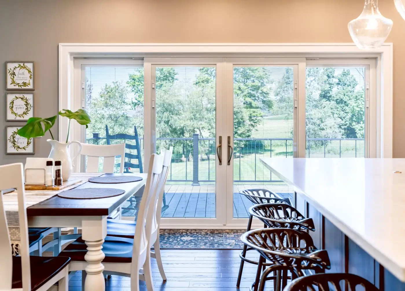 Inside a large open concept kitchen with a dark table and white chairs to the left and a white kitchen counter to the right with different dark chairs along it, and a large set of French patio doors with internal blinds in white and white framing with two large windows to the sides and a backyard deck beyond.