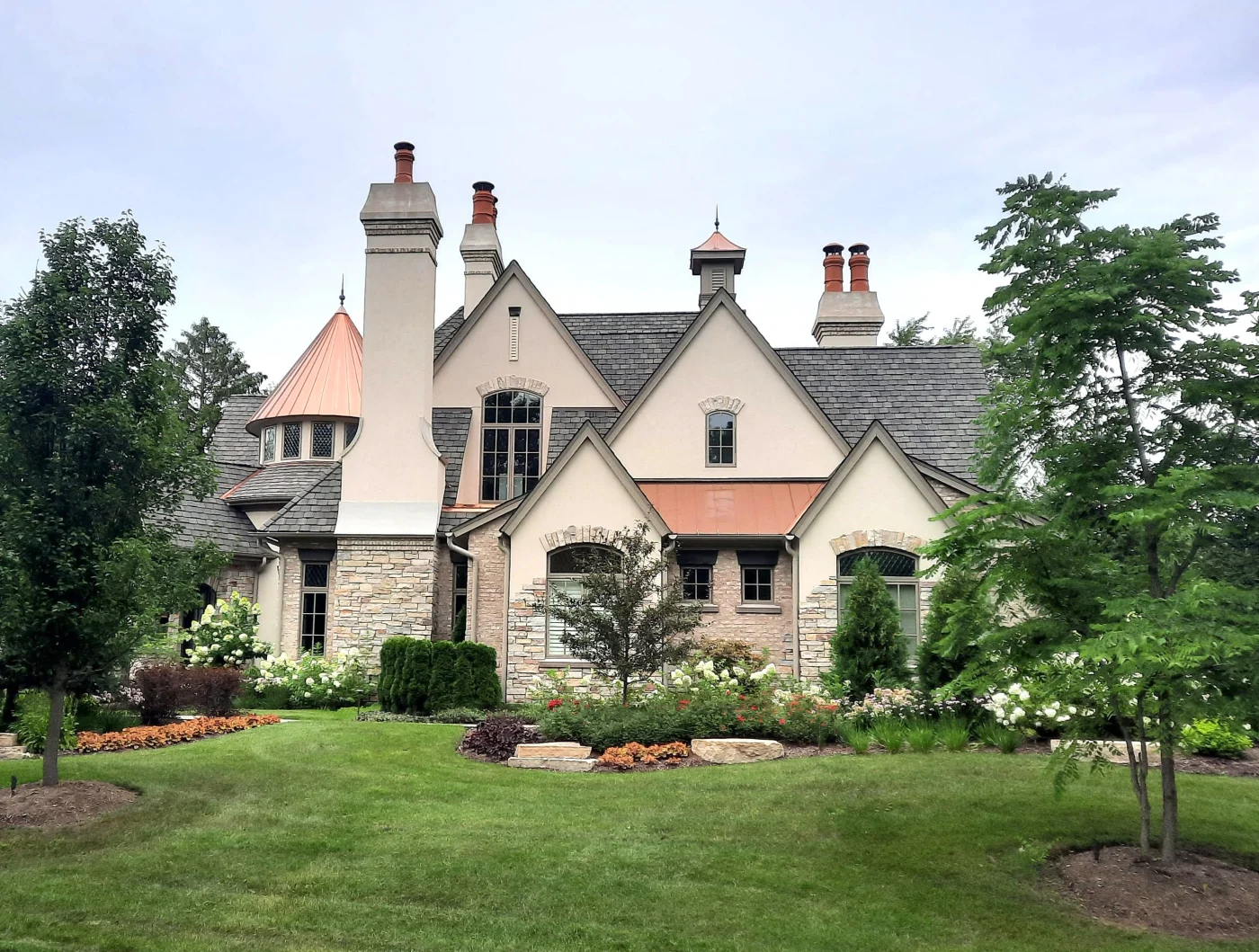 A very large luxurious home with a complex shingle roofing system with three chimneys and mainly engineered stone panel siding aside from stucco sections, various windows, and luxury landscaping out front with a manicured green lawn.