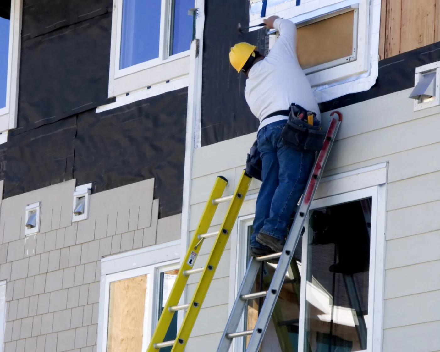 A siding installer wearing a yellow hard hat, white long sleeve shirt, jeans, and work shoes, up on a ladder holding a hammer, working on a section of siding around a window.