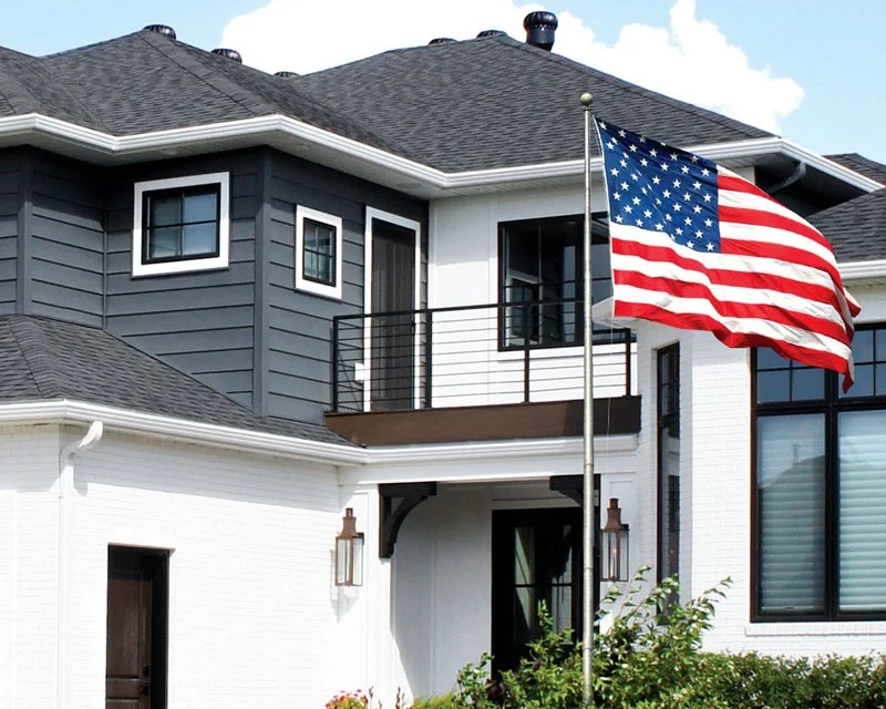 A large multi-level home with an American flag waving outside, a dark gray shingle roof, white window frames, dark windows, white gutters and soffit, and both white and dark gray steel siding.