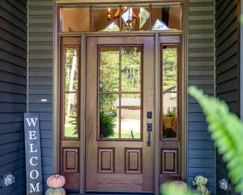 A beautiful single entry front door manufactured by ProVia with a dark cherry stain, matte black hardware, a large grid of four sections of clear glass, with a sidelite on both sides with the same glass, and a row of four small square picture windows above it.
