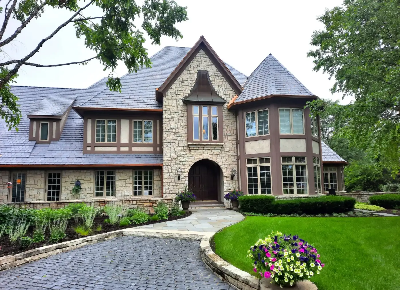 A large beautiful luxury home in the Chicagoland area with light brown stone panel siding, some stucco areas, darker brown trim, fifteen different sets of windows with traditional grids in light brown, along with large, thick wooden double entry doors without glass.