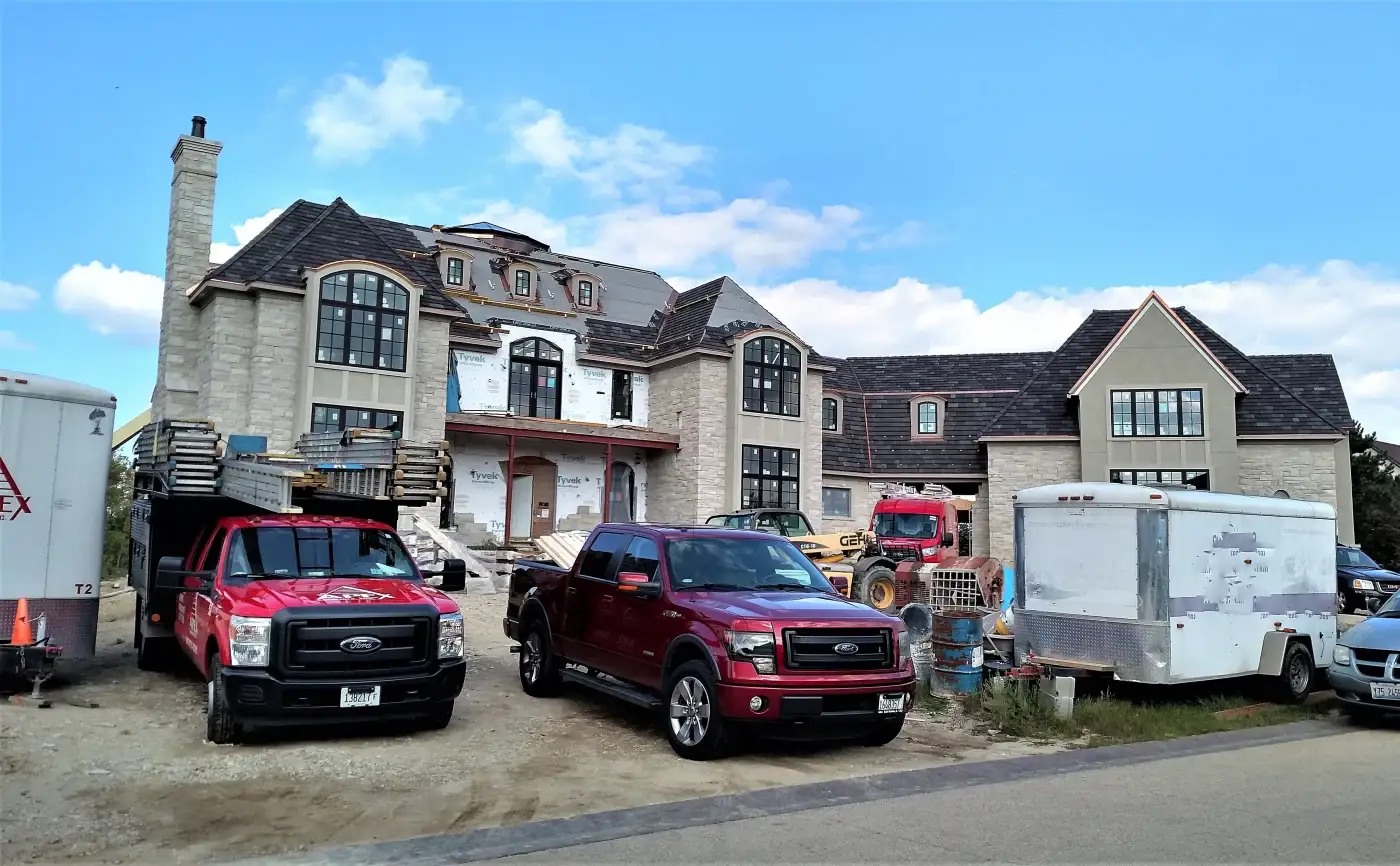 This is the construction site of a large luxury home in the Chicagoland area with new stone panel siding being installed, new premium high-end roofing being installed, and new recently-installed windows with the stickers still on them. Out front are a variety of vehicles including a white Apex Exteriors trailer and red truck.