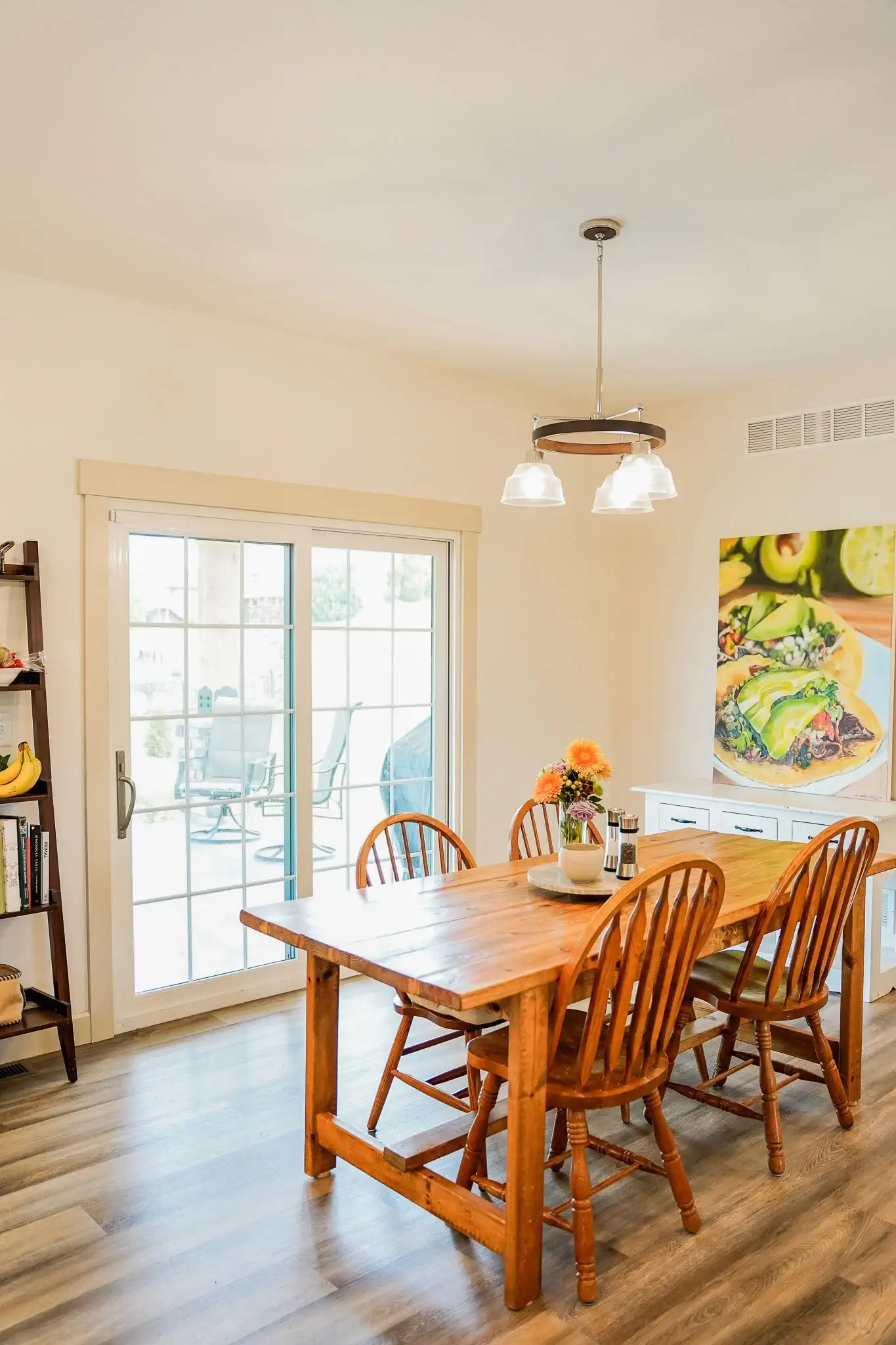 Comprehensive Patio Door Manufacturer Options For Chicagoland Homeowners A lovely dining room area in a home with a wooden table and four classic chairs, a painting with food, and a thick sliding glass patio door from ProVia with white colonial grids and bronze hardware.
