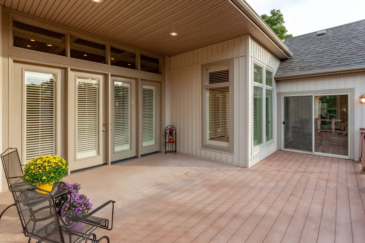 Patio Door Replacement Services In The Chicagoland Area A large back patio area with a wooden deck, two sets of patio doors, one of the left with internal blinds in the glass and two large windows on either sides of the doors, as well as a sliding glass patio door on the right. The siding and fascia are in nice panel siding with small light fixtures set within the covered area with soft brown gutters.
