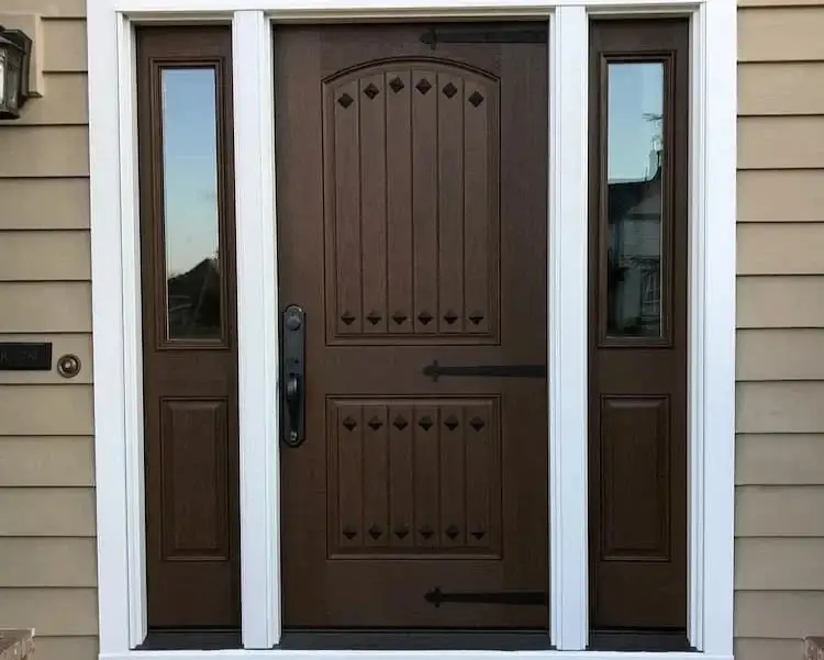 A stunning dark brown fiberglass entry door by Pella with no glass, but two sections of paneling with dark matte black hardware in it along with a black door handle, a half-glass sidelite on both sides, in a white frame.