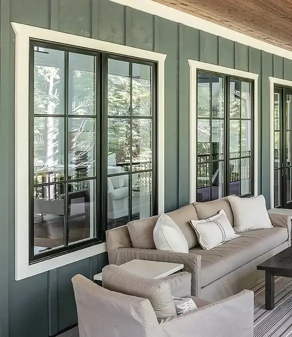 A set of three large casement windows with traditional grids and white framing seen on the outside of a residential covered porch with a couch, chair, coffee table, pillows, and a carpet.