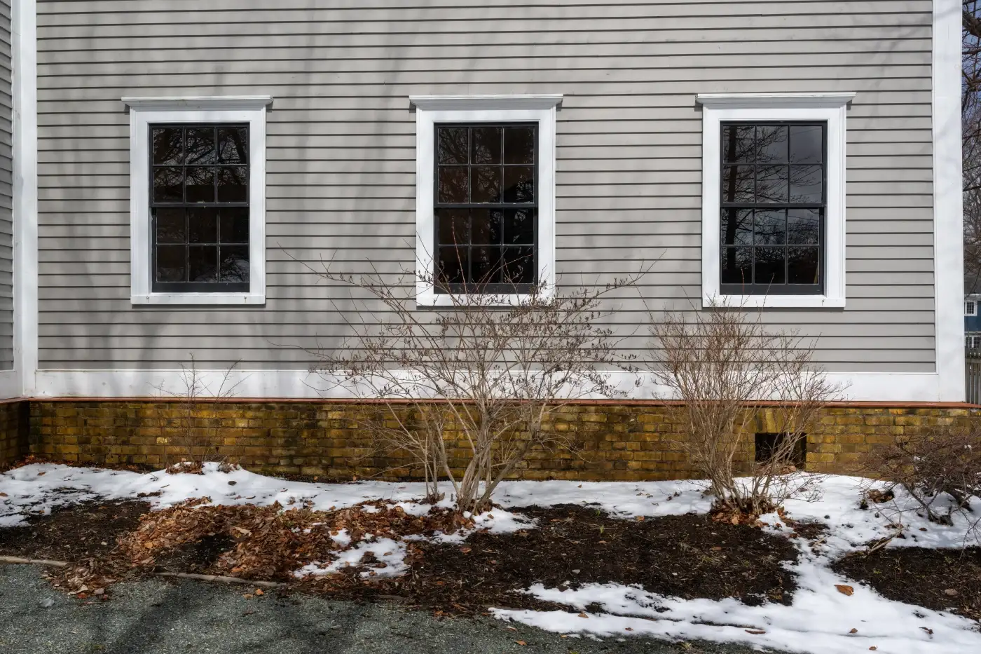 A set of three dark double-hung windows with white framing set in the side of a home with horizontal panel siding and white trim with a brick base. There’s some snow on the ground outside and the plants are barren of leaves.