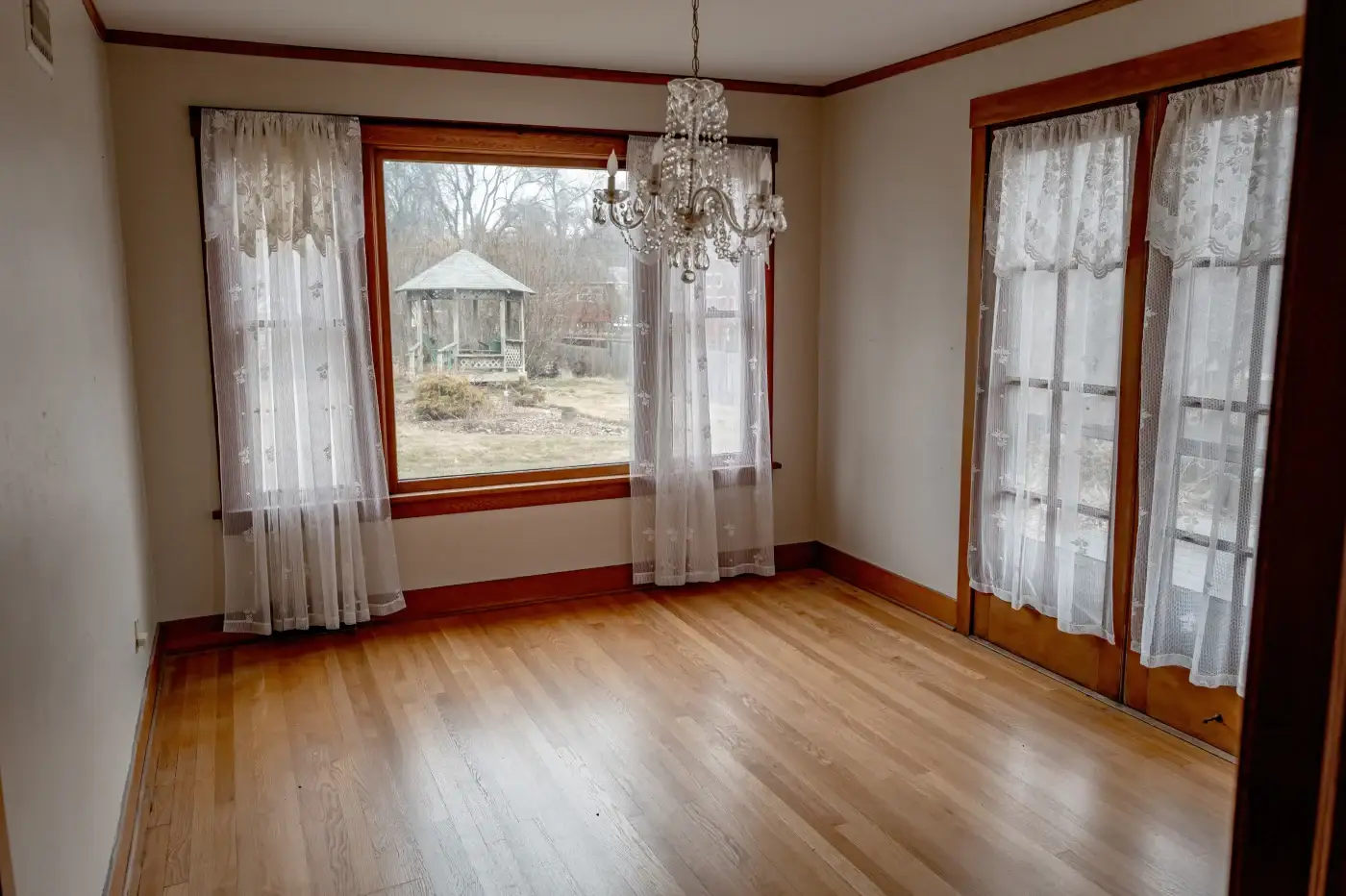 An empty residential room ready to be moved into, with a large picture window facing the backyard with double-hung windows to each side, wood framing, a wood panel floor, and an-entry door with a traditional glass grid to the other side of the room.