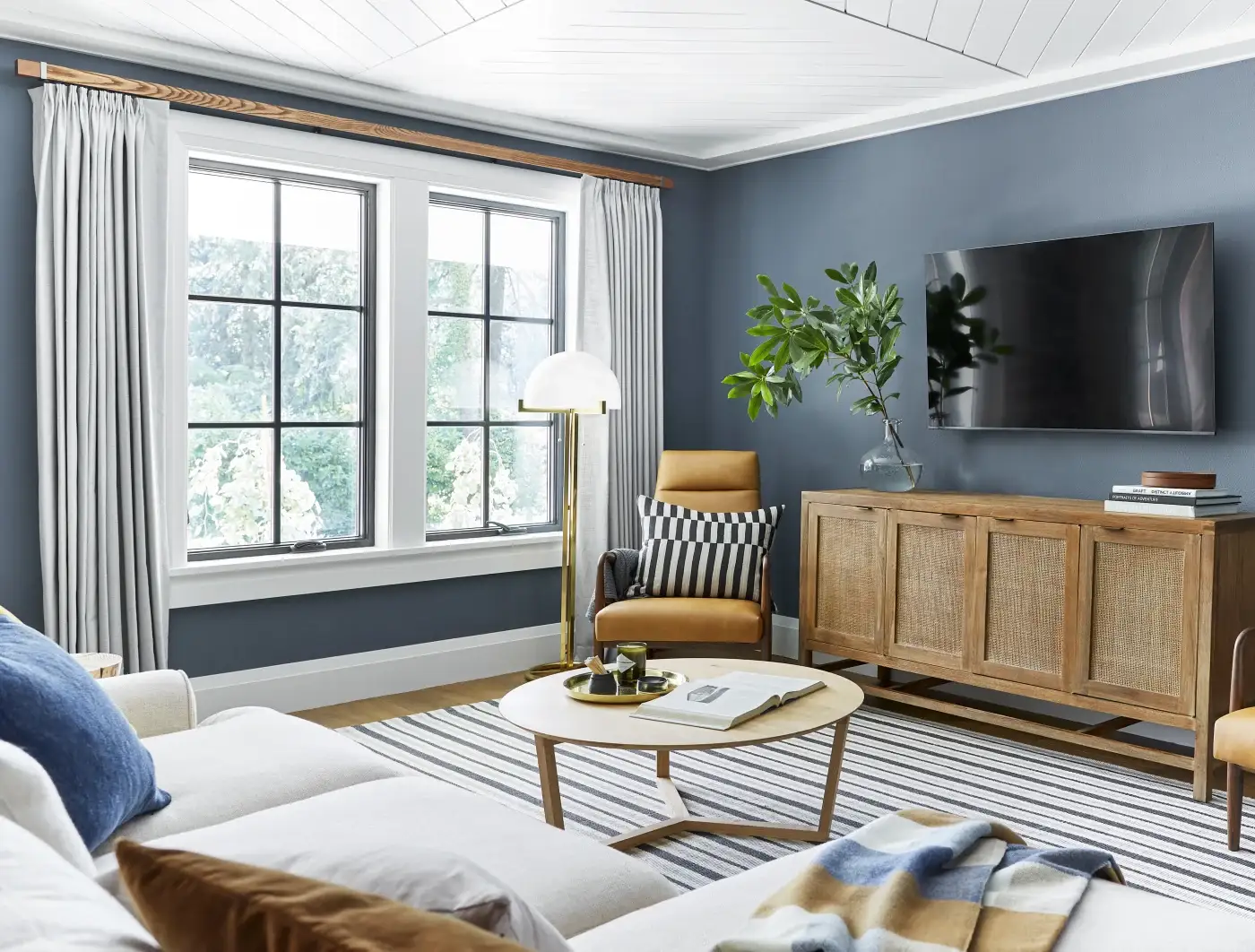 A lovely furnished living area with a big screen TV and TV table, different chairs and pillows, a large rug, a small coffee table, a tall lamp, and a set of large fiberglass windows with white framing and a darker internal grid.