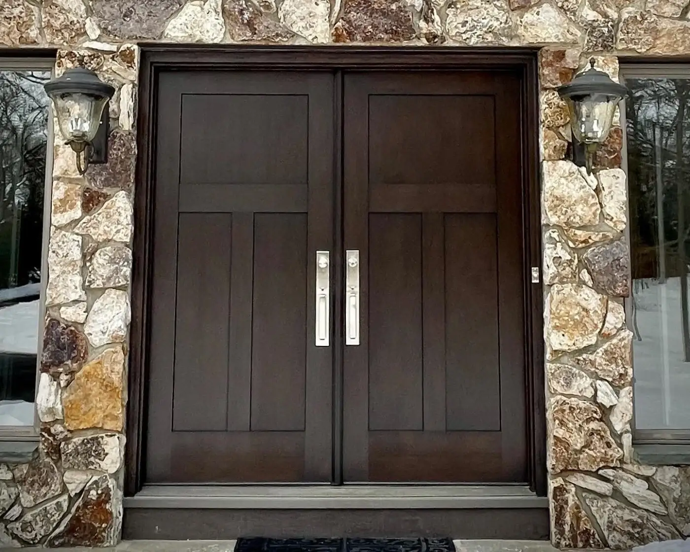 A large set of Therma-Tru double entry doors that are closed, with bright metal hardware and a small doorbell to the side, set within what looks like a stone veneer siding.