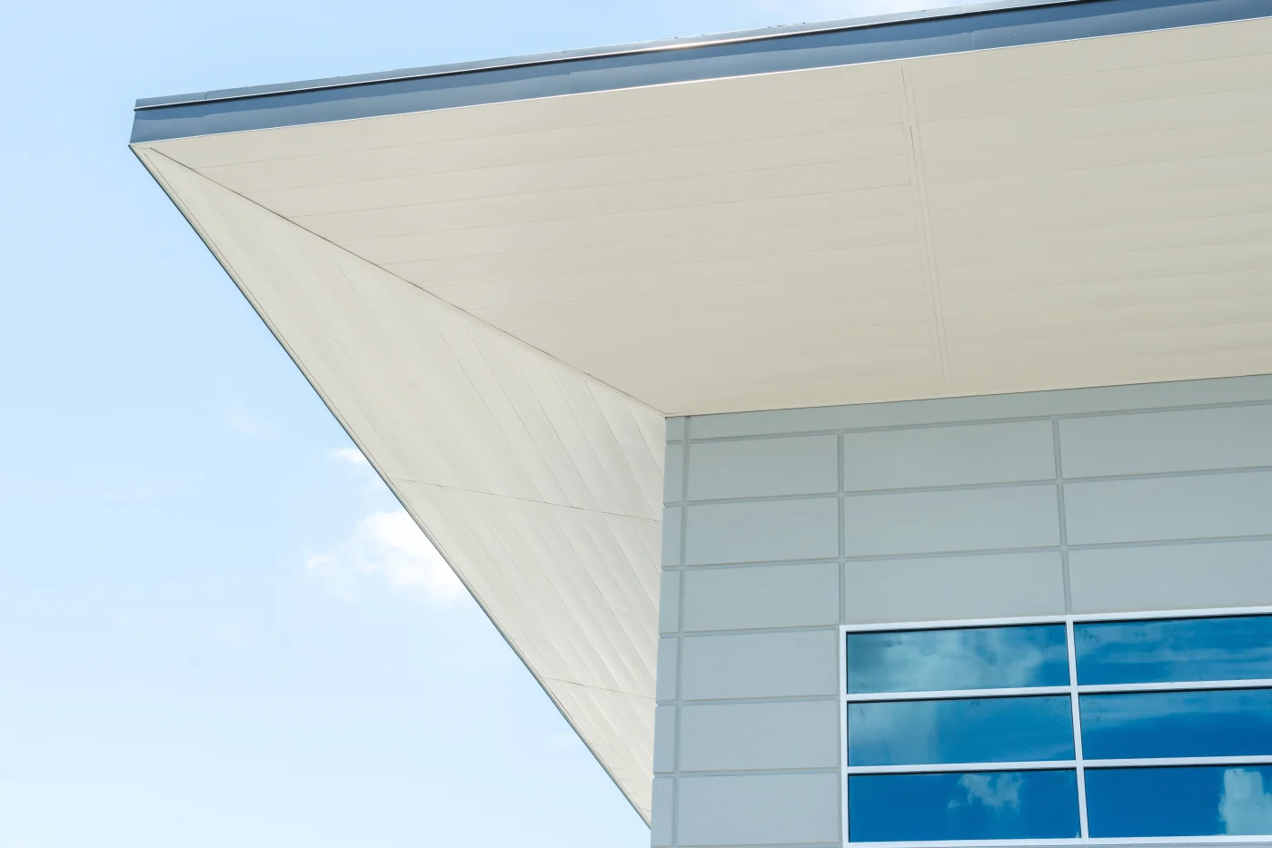 Chicagoland Commercial New Construction Soffit Services Close-up view of clean white commercial soffit panels installed beneath a sharply angled modern roof overhang, with light gray architectural wall panels and reflective blue-tinted windows, under a clear blue sky.
