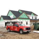 A new two-story custom home under construction in the Chicagoland area, partially wrapped in Tyvek and Kingspan house wrap. The black asphalt shingle roof has been installed, while the siding and trim are still unfinished. In front of the home is a bright red Apex Exteriors work truck with tool compartments and company branding, parked beside a large green construction dumpster on a gravel driveway.