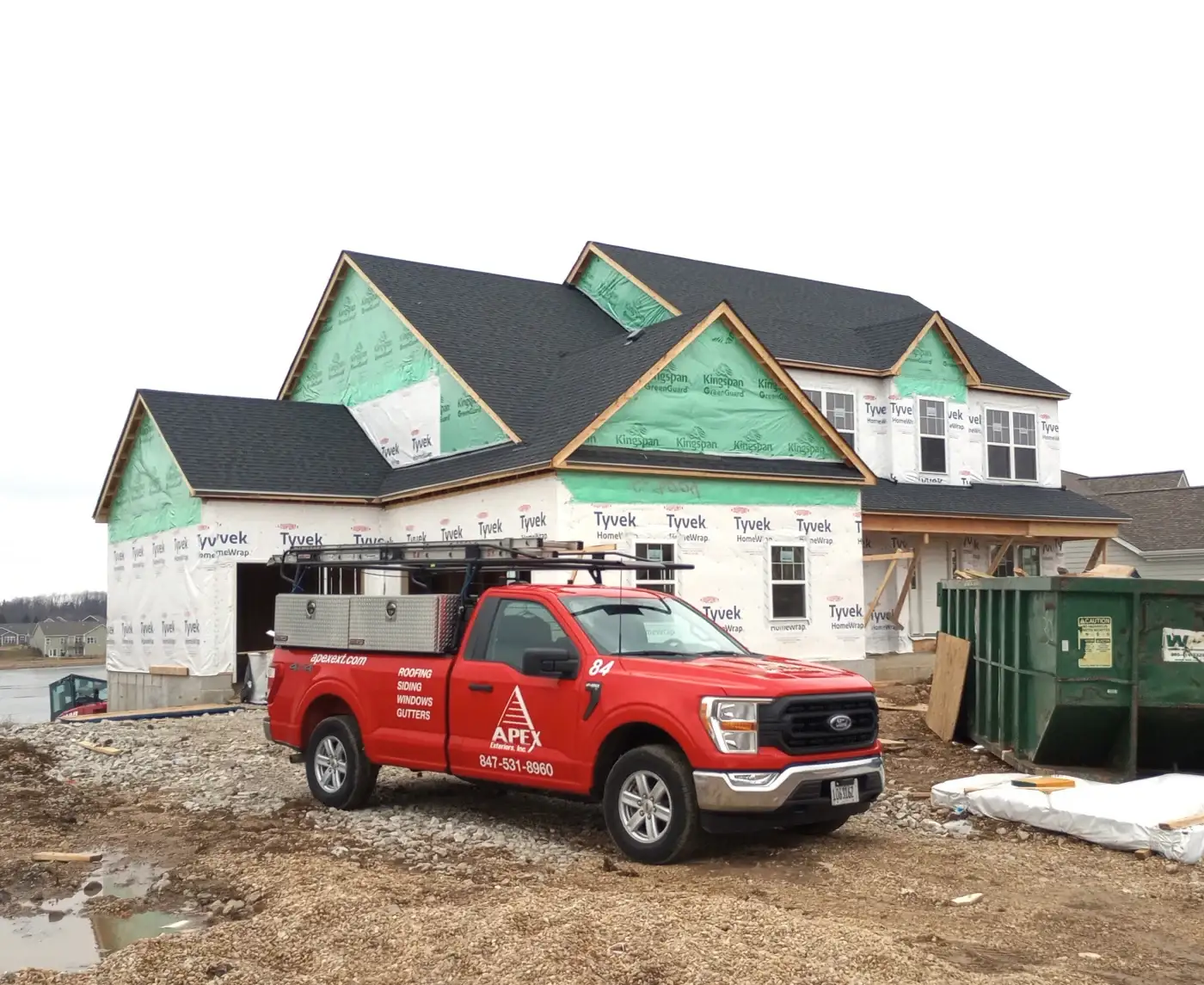 A new two-story custom home under construction in the Chicagoland area, partially wrapped in Tyvek and Kingspan house wrap. The black asphalt shingle roof has been installed, while the siding and trim are still unfinished. In front of the home is a bright red Apex Exteriors work truck with tool compartments and company branding, parked beside a large green construction dumpster on a gravel driveway.