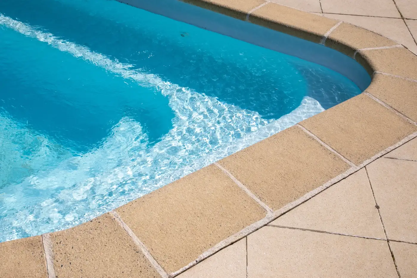 Customer-Centric Home Roofing Services In Chicagoland–Pool Deck Example Close-up view of a clean, inviting backyard pool with bright blue water and tan concrete coping. The pool is surrounded by light-colored tile decking, showing a well-maintained residential outdoor space under clear sunlight.