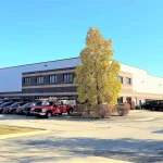 A bright, clear day outside the Apex Exteriors headquarters in South Elgin, Illinois. The two-story brick-and-metal building is surrounded by neatly maintained trees and landscaping in full autumn color. A long row of red Apex-branded trucks is lined up in the parking lot, showcasing the company’s readiness and strong fleet presence.