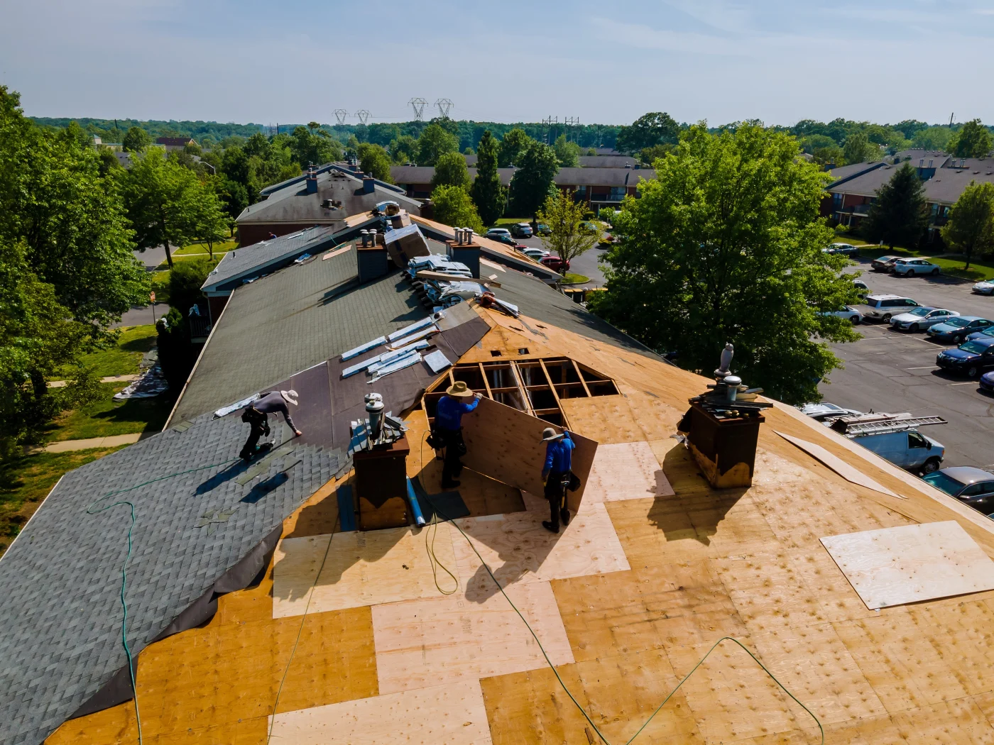 A team of roofing professionals working on a multi-family roof replacement project. The image shows old shingles being removed and new underlayment and decking being installed. The surrounding apartment complex and full parking lot highlight the occupied nature of the building during the re-roofing process.