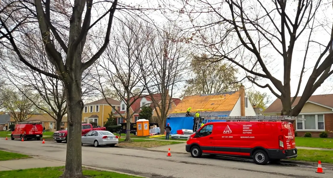 An Apex Exteriors project manager checks on a residential roofing crew in the Chicagoland area. Workers on the roof are wearing high-visibility safety gear and are properly secured with ropes and fall protection. Bright red Apex service vans are parked along the street, with safety cones in place. The home under construction has exposed roof decking, and job materials are staged on the lawn, showcasing an organized and compliant worksite.