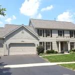 A two-story home in the Chicagoland area featuring a completed CertainTeed asphalt shingle roof in a light gray tone. The home has beige James Hardie siding, dark brown shutters, and a three-car garage. A manicured lawn, shrubs, and trees frame the property under a bright, sunny sky with scattered clouds.
