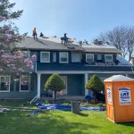An Apex Exteriors crew works on a residential roofing project in the Chicagoland area, visibly following OSHA safety guidelines. Multiple workers are secured with safety harnesses and ropes while removing old roofing materials and preparing the roof deck. The home is a blue two-story house with white trim, surrounded by springtime landscaping and a blooming magnolia tree.
