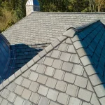A newly installed roof featuring CertainTeed Grand Manor shingles on a large, elegant home. The architectural shingles display a deep, dimensional slate look with varied tones of gray. The roof’s complex angles, hip ridges, and steep slopes highlight the shingles’ layered design. In the background, turrets and stone chimney details add to the upscale aesthetic, surrounded by leafy trees under clear daylight.
