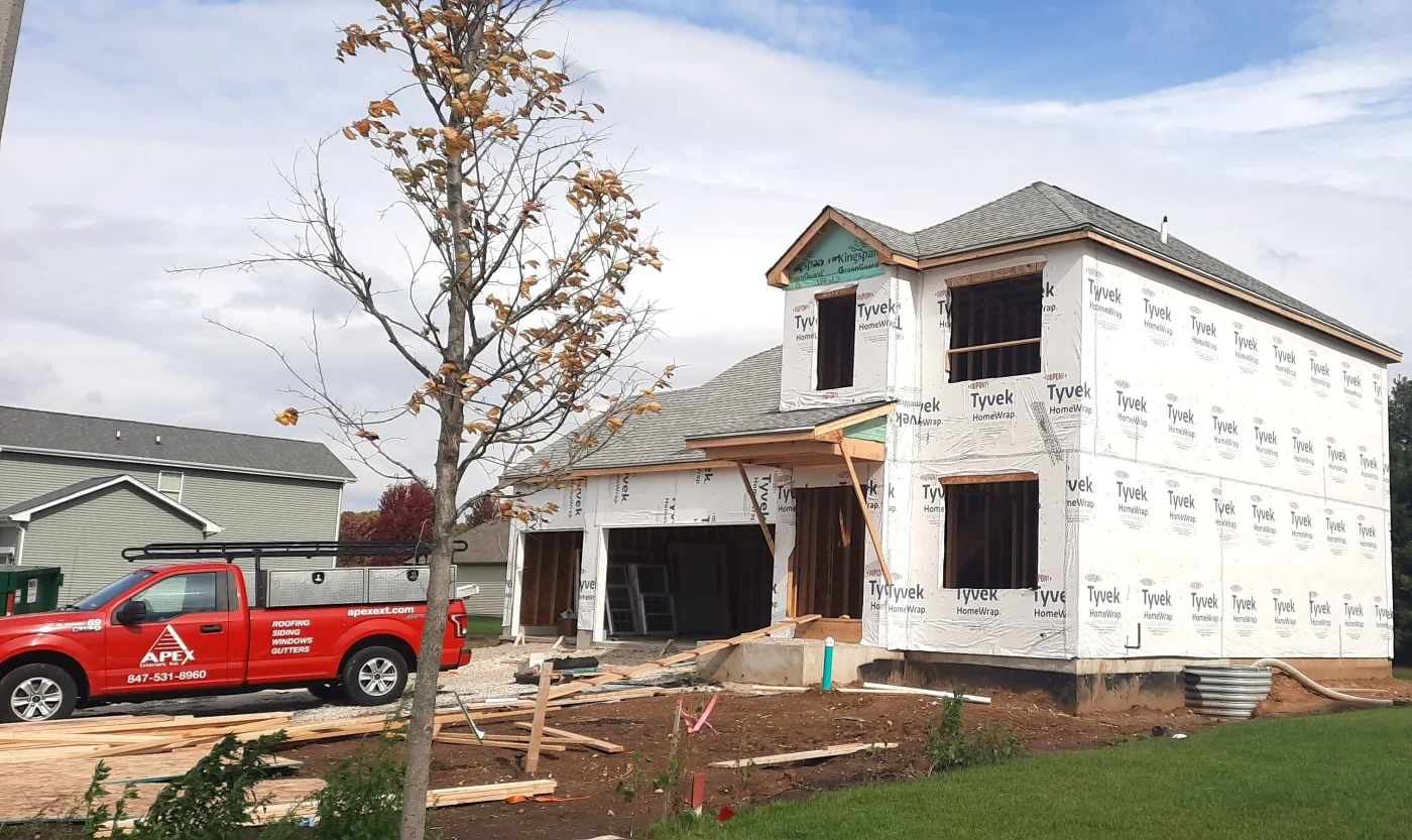 Outside a new construction site of a regular Chicagoland home, with the frame built and wrapped in Tyvek house wrap, showing where the windows, front entry, and garage doors will go, with a new shingle roofing installed by Apex Exteriors, with one of their red work trucks parked in what will become the driveway.