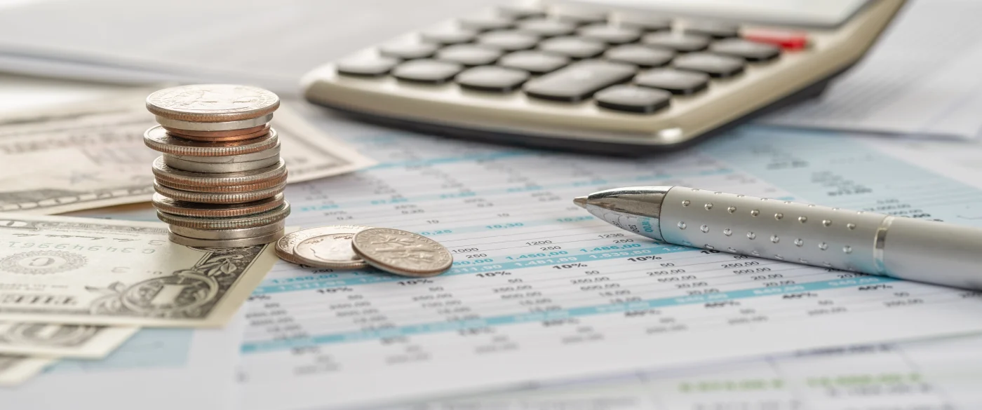 Some American money and coins atop some financial paperwork with a pen and a calculator on a table.