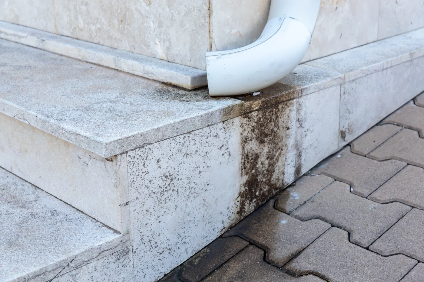 The bottom of an incorrectly placed gutter downspout causing ugly water damage and streaking on some concrete steps and stone pathway.