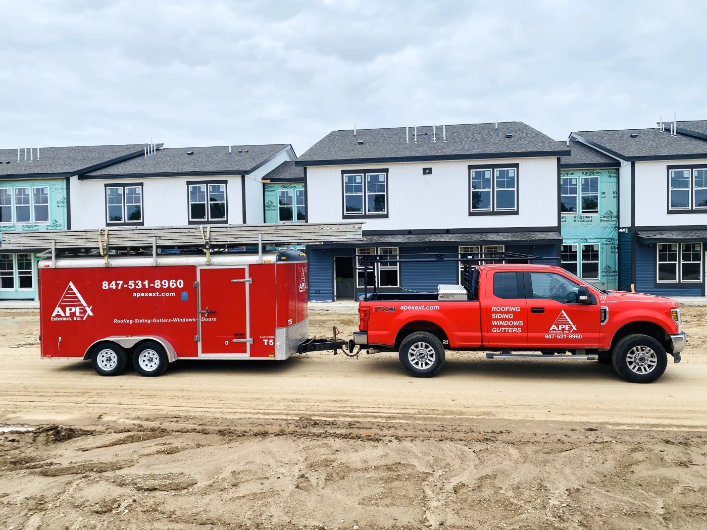 An Apex Exteriors work truck and enclosed trailer parked at a multi-family construction site in the Chicagoland area. New townhomes are partially completed, featuring fresh siding, installed windows, and roofing work in progress. The clean job site and branded equipment reflect Apex’s full-service capabilities and professional approach.