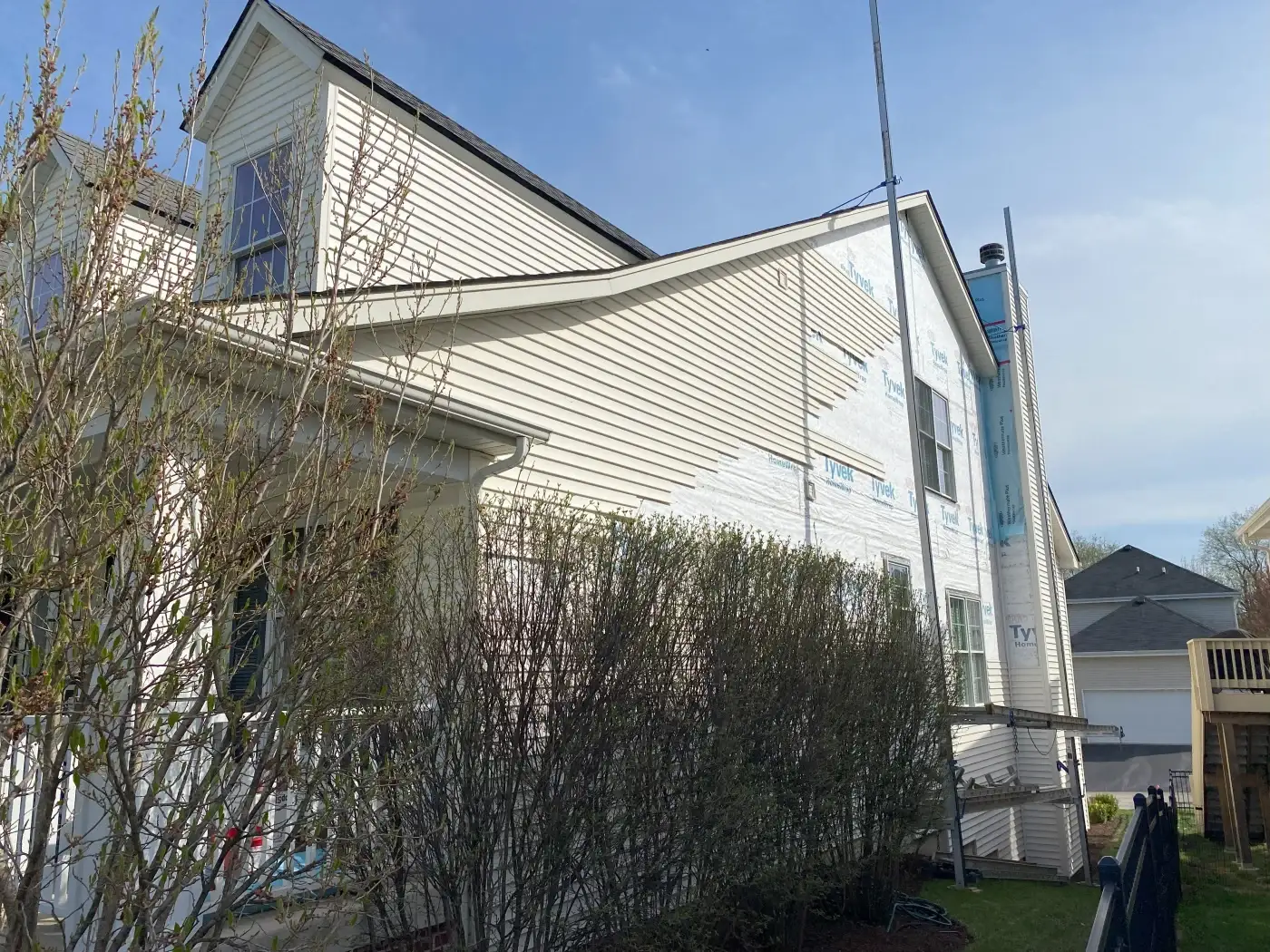 A two-story suburban home is undergoing siding replacement. The left half of the house already has new beige siding, while the right side remains covered in white house wrap. A mechanical scaffolding system is set up vertically along the house’s right side. In the foreground, neatly trimmed bushes line the side of the home. The weather is clear, with a bright blue sky and no visible clouds. The overall setting appears to be a quiet residential neighborhood.