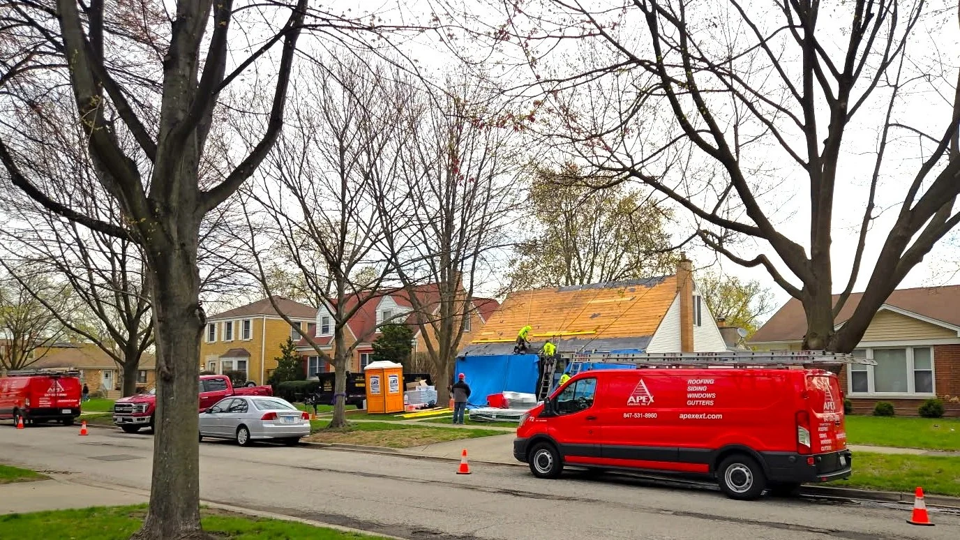 An Apex Exteriors red work van sits on the road outside a home having it’s roof replaced by Apex roofing teams, showing them using safety harnesses and ropes, being watched by a crew leader in a red hard hat.