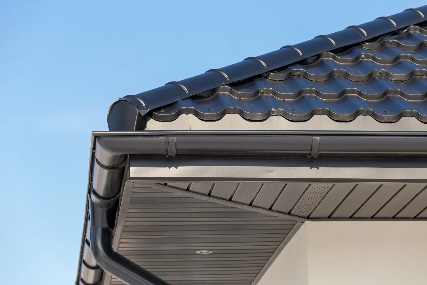 Corner Of The New Modern House With Gutter Roof And Wall A close-up view of a building’s roof overhang, showing dark perforated soffit panels and matching fascia beneath a black tiled roof. The clean lines and precise installation highlight proper soffit ventilation and trim detailing under a clear blue sky.