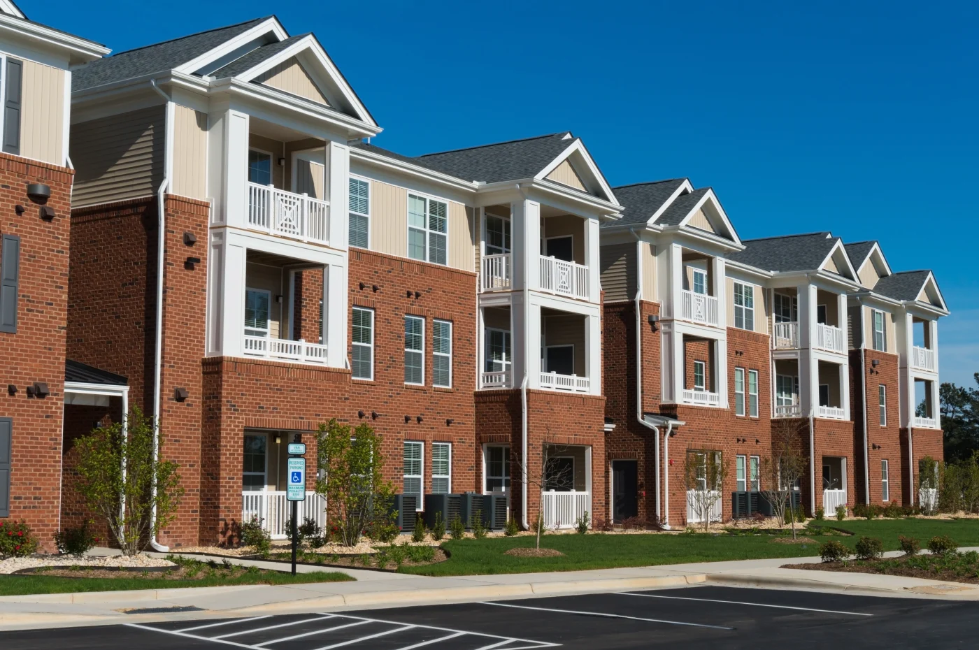 A newly constructed three-story apartment building featuring a mix of brick and light siding with clean, architectural roofing. The paved parking lot is freshly striped and landscaped, reflecting a completed new construction project. The building's crisp lines and modern materials emphasize quality exterior and roofing installation.
