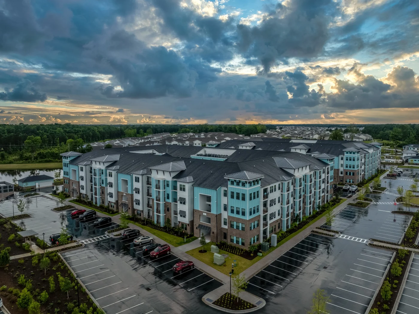A drone view of a modern multi-story apartment complex with dark gray roofing and teal and white exterior paneling. The buildings are surrounded by wet pavement and overcast skies, suggesting recent rain. The large-scale layout highlights the roofing system installed across the expansive multi-family property.
