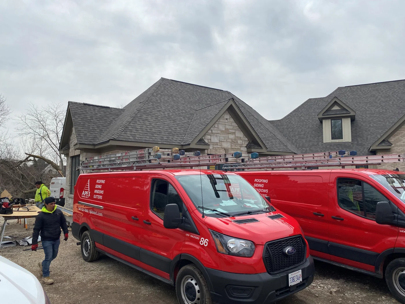 Two red Apex Exteriors work vans outside on a new home construction site in the Chicagoland area, with the new home behind them showing the new roofing system and stone tile panel siding system on a cloudy day.