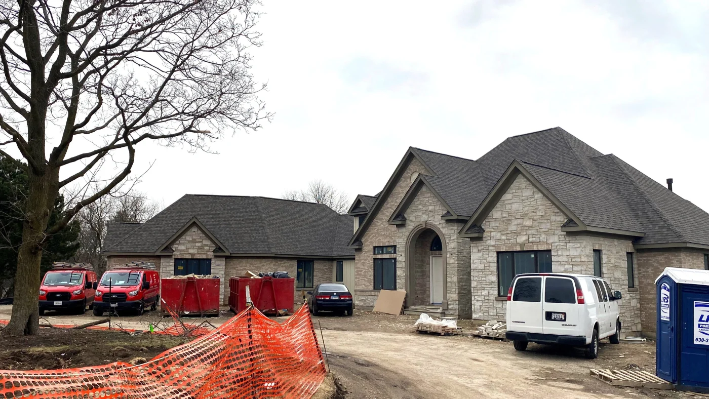 Apex Exteriors Uses An Old-School, Classic Kind Of Professionalism Outside a new construction site of a large luxury home in the Chicagoland area, showing a large, single-story structure with newgray stone siding and dark gray shingle roofing system. There are Apex Exteriors work vans parked outside it with dumpsters and a porta potty set up.