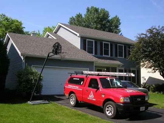 Apex Exteriors Sends Fully Prepared Roofing Services Consultants An Apex Exteriors red work truck outside a quaint Chicagoland home with a white garage and trim, four single-hung windows upstairs, a freshly-cut green lawn, and a new roofing shingle roofing system.