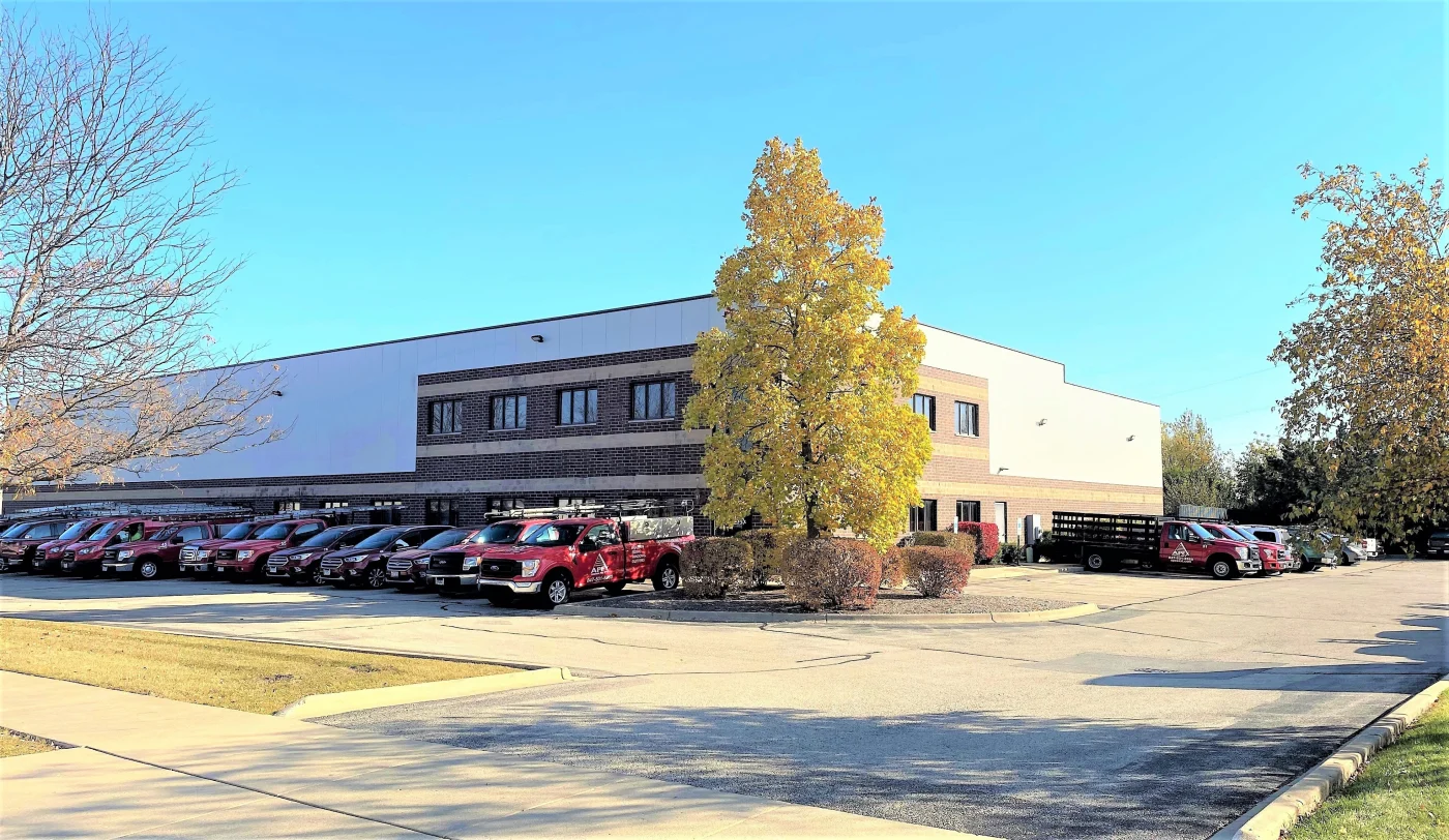Apex Exteriors’ South Elgin headquarters features a modern brick building with a full fleet of red company trucks neatly lined up in front. The well-maintained lot, seasonal trees, and clear blue sky reflect the company’s professional, established presence.