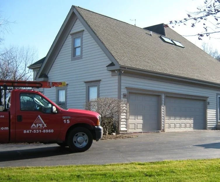 Apex Exteriors Specialized Roofing Service Consultants Chicagoland The front end of an Apex Exteriors work truck outside a Chicagoland home with a three-car garage, light brown panel siding, brown gutters, dark brown trim, and an asphalt shingle roofing system.