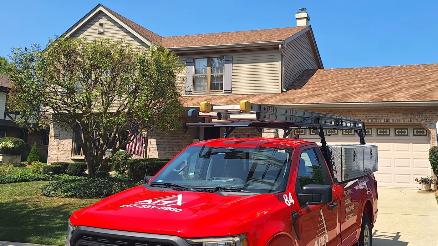 A nice single-family split-level home in the Chicagoland area from the street with an Apex Exteriors work truck in the driveway. The home has stone siding around the main section, with light brown panel siding upstairs, new brown asphalt shingle roofing with light brown fascia, gutters, and trim.