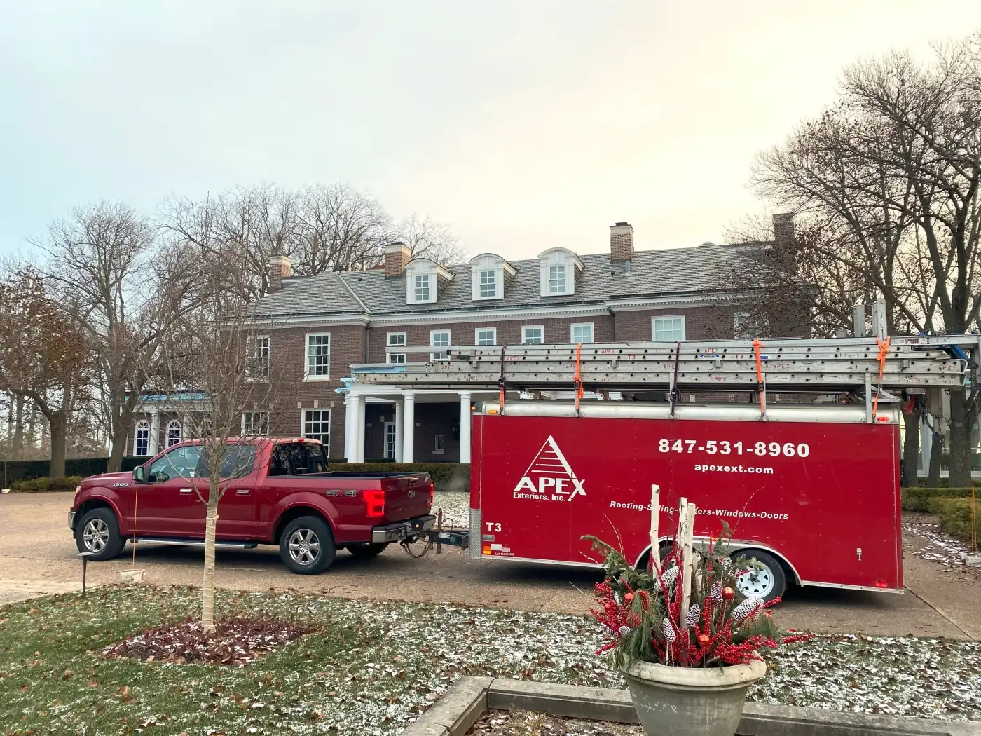 A large, long two-story brick home with fourteen visible windows with white framing and grids, white specialty moulding around the roof, four chimneys, a regal covered entrance with white pillars, and a black work truck sitting in the driveway out front pulling a red Apex Exteriors trailer with a variety of ladders stacked on top of it.
